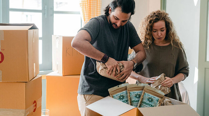 man and a woman packing kratom and other things inside a box near other boxes indoors