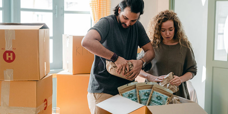 man and a woman packing kratom and other things inside a box near other boxes indoors