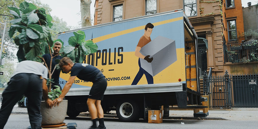 three men carrying a big plant in a pot and a hauling truck and apartment building in the background